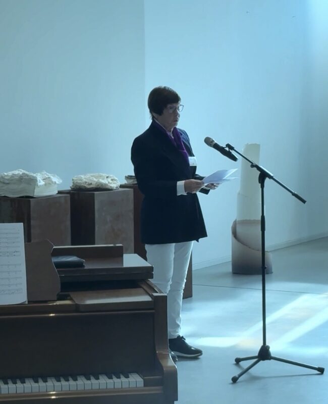 Eine Person mit Brille, dunklem Blazer, weißer Hose und violettem Schal steht an einem Mikrofon und liest von Papieren ab. Im Vordergrund steht ein Klavier mit Notenblättern. Im Hintergrund sind bei hellem, natürlichem Licht Skulpturen und Kunstwerke zu sehen.