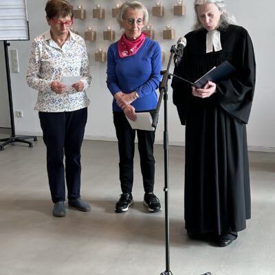 Drei ältere Frauen stehen in einem Raum vor einem Mikrofon. Zwei tragen Freizeitkleidung und Schals und halten Papiere in der Hand. Die dritte trägt ein schwarzes Gewand, hält ebenfalls Papiere in der Hand und scheint eine Zeremonie zu leiten. Hinter ihnen stehen viele brennende Kerzen auf einem Wanddisplay.