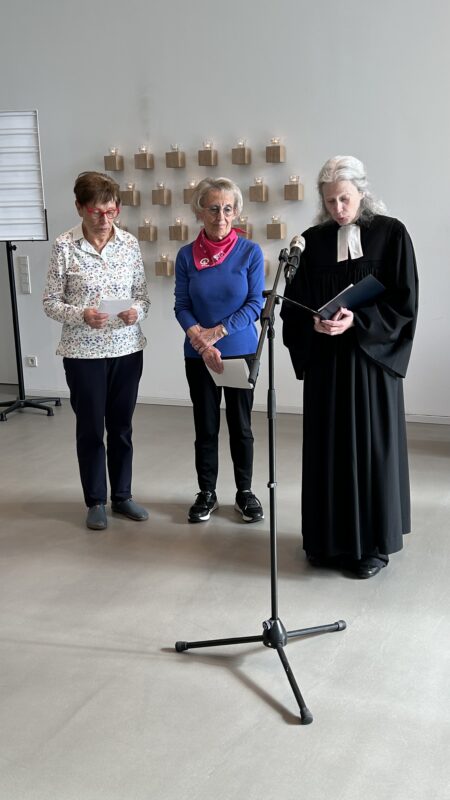 Drei ältere Frauen stehen in einem Raum vor einem Mikrofon. Zwei tragen Freizeitkleidung und Schals und halten Papiere in der Hand. Die dritte trägt ein schwarzes Gewand, hält ebenfalls Papiere in der Hand und scheint eine Zeremonie zu leiten. Hinter ihnen stehen viele brennende Kerzen auf einem Wanddisplay.