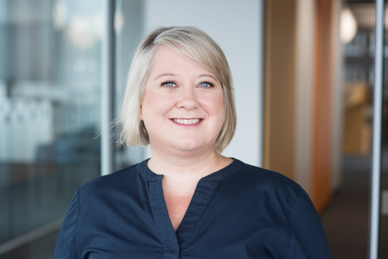 Eine lächelnde Frau mit kurzen blonden Haaren und heller Haut trägt eine marineblaue Bluse. Sie steht in einem modernen, hell erleuchteten Büroflur mit Glaswänden und unscharfen Hintergrundelementen.