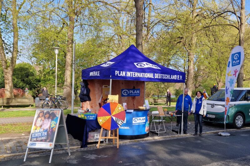 Ein Stand von Plan International Deutschland e.V. mit einem blauen Vordach steht im Freien zwischen Bäumen. Vorne steht ein buntes Glücksrad, daneben stehen zwei Personen. Rund um den Stand sind Plakate und Banner mit Kinderthemen und dem Plan-Logo angebracht.