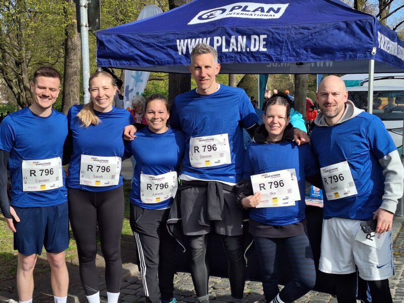 Sechs Personen in blauen Hemden und mit Startnummern stehen dicht beieinander und lächeln in die Kamera. Sie befinden sich im Freien, unter einem blauen Zelt mit der Aufschrift PLAN.DE. Im Hintergrund sind Sonnenlicht und Bäume zu sehen, was auf eine lustige, aktive Veranstaltung hindeutet.