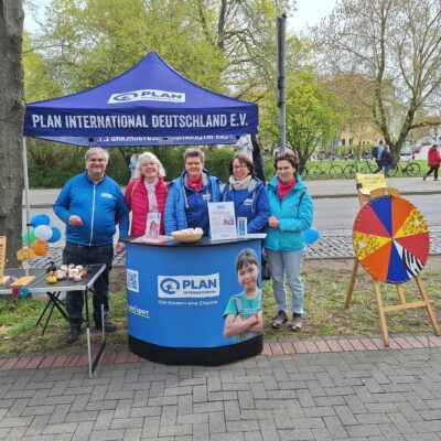 Fünf Menschen stehen lächelnd unter einem blauen Zelt von Plan International Deutschland e.V. an einem Stand im Freien. Auf dem Tisch liegen Broschüren, eine Spendenbox und das Foto eines Mädchens. In der Nähe stehen ein Spinnrad und ein Holzspiel. Im Hintergrund sind Bäume und eine Straße zu sehen.