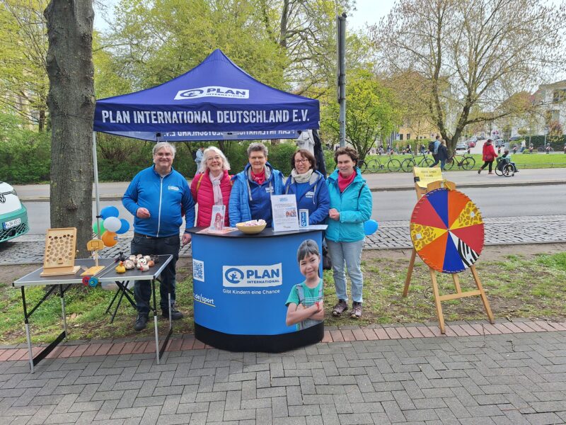 Fünf Menschen stehen lächelnd unter einem blauen Zelt von Plan International Deutschland e.V. an einem Stand im Freien. Auf dem Tisch liegen Broschüren, eine Spendenbox und das Foto eines Mädchens. In der Nähe stehen ein Spinnrad und ein Holzspiel. Im Hintergrund sind Bäume und eine Straße zu sehen.