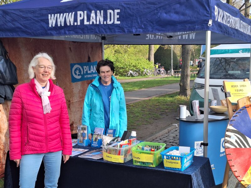 Zwei Frauen stehen an einem Infostand im Freien unter einem blauen PLAN.DE-Vordach. Auf dem Tisch liegen Broschüren, Körbe und ein buntes Preisrad. Eine Frau trägt eine rosa Jacke und eine Brille, die andere trägt blau. Im Hintergrund sind Menschen und Grünflächen zu sehen.