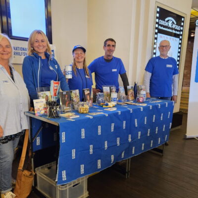 Fünf Personen stehen hinter einem Tisch, der mit einem blauen Tuch von Plan International bedeckt ist, und legen Broschüren und Werbeartikel aus. Alle tragen blaue Hemden oder Jacken mit dem Plan-Logo. Rechts ist ein großes Banner von Plan International zu sehen, und im Hintergrund ist eine Treppe zu erkennen.