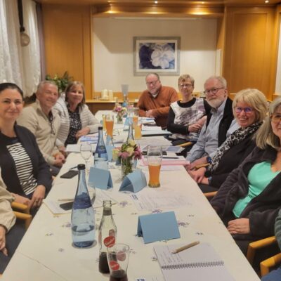 Zwölf Erwachsene, Männer und Frauen, sitzen um einen langen Tisch mit Getränken, Notizbüchern und Blumen in einem warm beleuchteten Raum. Die meisten lächeln in die Kamera, was auf ein freundliches Treffen oder eine Zusammenkunft schließen lässt. Die Atmosphäre wirkt ungezwungen und einladend.