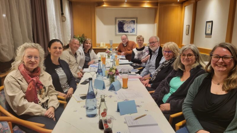 Zwölf Erwachsene, Männer und Frauen, sitzen um einen langen Tisch mit Getränken, Notizbüchern und Blumen in einem warm beleuchteten Raum. Die meisten lächeln in die Kamera, was auf ein freundliches Treffen oder eine Zusammenkunft schließen lässt. Die Atmosphäre wirkt ungezwungen und einladend.