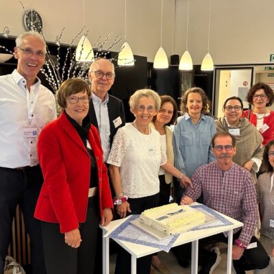 Eine Gruppe von zehn Erwachsenen versammelt sich lächelnd um einen rechteckigen Kuchen auf einem kleinen Tisch in einem hellen Innenraum. Einige tragen Namensschilder. Darüber hängen Hängelampen, und im Hintergrund sind Regale, eine Uhr und Informationsplakate zu sehen.