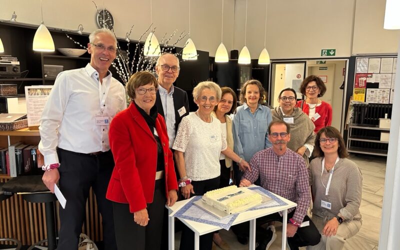 Eine Gruppe von zehn Erwachsenen versammelt sich lächelnd um einen rechteckigen Kuchen auf einem kleinen Tisch in einem hellen Innenraum. Einige tragen Namensschilder. Darüber hängen Hängelampen, und im Hintergrund sind Regale, eine Uhr und Informationsplakate zu sehen.