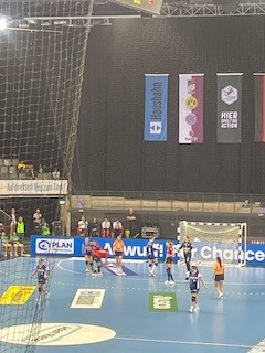 Handballspiel in der Halle; blau-weiße Spieler verteidigen gegen eine orangefarbene Mannschaft in Tornähe. Dahinter hängen Transparente an einer schwarzen Wand. Im Vordergrund ist ein Netz zu sehen, und auf der linken Seite sitzen einige Zuschauer auf der Tribüne.