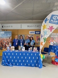 Vier Personen stehen hinter einem Tisch, der mit einem blauen Tuch mit dem Logo von Plan International bedeckt ist. Auf dem Tisch liegen Broschüren und Luftballons. Hinter ihnen sind Plan-Banner und eine Fahne zu sehen. Der Schauplatz scheint eine überdachte Veranstaltungshalle mit Schildern für Blöcke und WC zu sein.