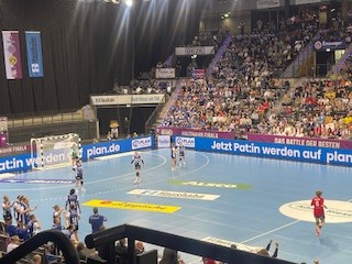 Hallenhandballspiel in einer vollen Halle. Eine Mannschaft in Weiß und Blau steht in der Nähe des Tores, während ein Spieler in Rot auf der rechten Seite mitten im Spiel ist. Die Zuschauer füllen die Tribünen; Banner und Werbung säumen den Spielfeldrand.