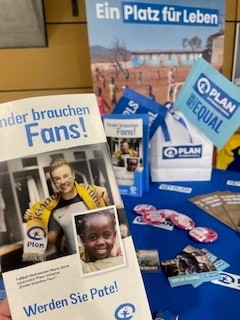 Eine Hand hält ein deutsches Flugblatt, das zwei lächelnde Kinder zeigt, von denen eines einen Fußballschal trägt, vor einem blauen Plan International-Display mit Flugblättern, Fahnen, Buttons und dem im Hintergrund sichtbaren Slogan "Ein Platz für Leben".