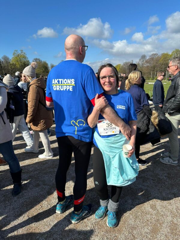 Eine lächelnde Frau mit Brille, blauem Hemd und Startnummer 38 steht neben einem glatzköpfigen Mann in einem blauen AKTIONS GRUPPE-Hemd, der seinen Arm um sie gelegt hat. Sie befinden sich an einem sonnigen Tag im Freien in einer Menschenmenge, mit Bäumen und Wolken im Hintergrund.
