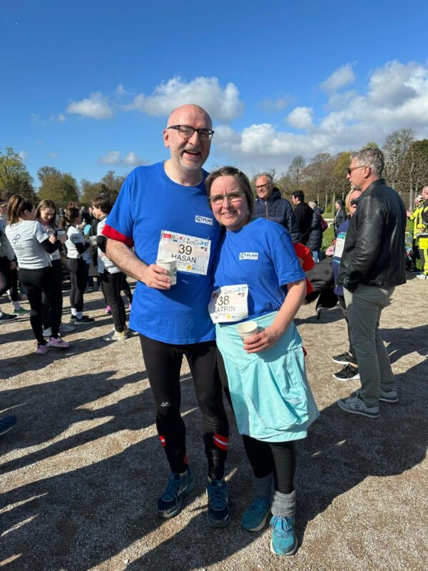 Ein lächelnder Mann und eine lächelnde Frau stehen bei einer Laufveranstaltung im Freien zusammen, beide tragen blaue Hemden und Startnummern. Sie posieren für die Kamera mit anderen sportlich gekleideten Personen im Hintergrund, unter einem sonnigen Himmel mit Wolkenfeldern und grünen Bäumen.