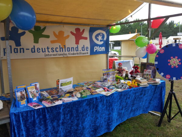 Ein farbenfroher Stand im Freien mit blauer Tischdecke bietet Bücher, Spielzeug und Flyer an. Luftballons schmücken das Zelt; auf einem Transparent steht plan-deutschland.de Internationales Kinderhilfswerk. Rechts steht ein sich drehendes Preisrad. Die Szene deutet auf ein Volksfest oder eine Wohltätigkeitsveranstaltung hin.