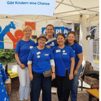 Fünf Frauen in blauen "Plan International"-Shirts stehen lächelnd unter einem Zelt bei einer Freiluftveranstaltung. Über ihnen prangt ein Banner mit der Aufschrift "PLAN International - Gibt Kindern eine Chance". Sie scheinen Freiwillige zu sein und stehen dicht beieinander.