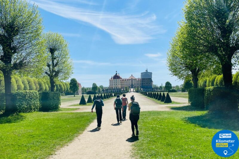 Vier Personen gehen auf einem sonnenbeschienenen Kiesweg, der von gepflegten Bäumen gesäumt ist, auf ein großes schlossähnliches Gebäude unter blauem Himmel zu. Der üppige grüne Rasen und die Hecken rahmen die Szene ein. In der unteren rechten Ecke befindet sich ein blaues "Kilometerzähler"-Abzeichen.