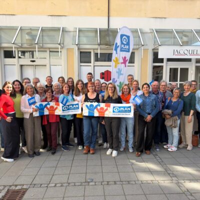 Eine Gruppe von etwa 30 lächelnden Menschen posiert vor einem Gebäude namens "JACQUELINE". Einige halten blaue und orangefarbene "Plan International"-Schilder und Transparente, um ihre Unterstützung für die Organisation zu zeigen. Die Umgebung ist hell mit Sonnenlicht und einem gepflasterten Bürgersteig.