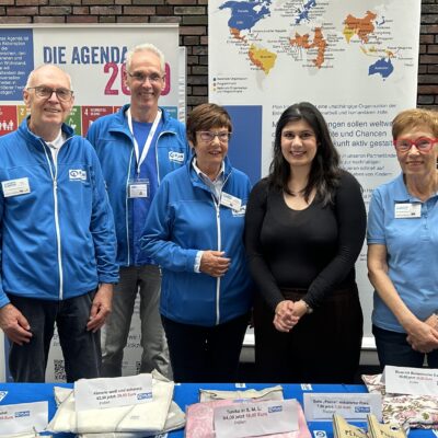 Fünf Personen stehen hinter einem Ausstellungstisch mit Broschüren, Textilien und Kunsthandwerk. Sie tragen blaue Jacken, außer einer Frau in Schwarz. Hinter ihnen befinden sich Plakate mit Landkarten, Text und einem runden farbigen Logo. Der Hintergrund ist eine Wand aus rötlich-braunen Ziegeln.