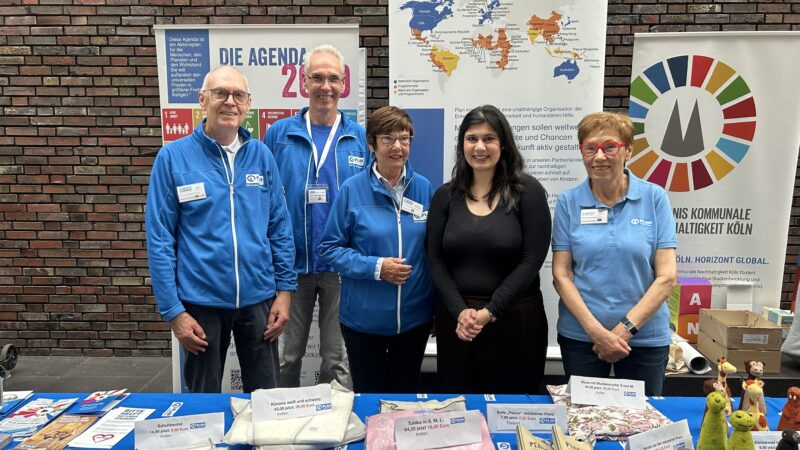 Fünf Personen stehen hinter einem Ausstellungstisch mit Broschüren, Textilien und Kunsthandwerk. Sie tragen blaue Jacken, außer einer Frau in Schwarz. Hinter ihnen befinden sich Plakate mit Landkarten, Text und einem runden farbigen Logo. Der Hintergrund ist eine Wand aus rötlich-braunen Ziegeln.