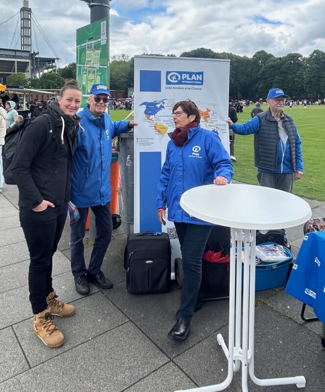 Vier Personen stehen an einem runden Tisch und einem Stand der Organisation PLAN im Freien. Hinter ihnen befinden sich eine Kartenanzeige, Menschen und eine Wiese. Drei von ihnen tragen blaue PLAN-Jacken und Hüte; einer trägt schwarze Kleidung und hellbraune Stiefel. Es ist bewölkt und locker.