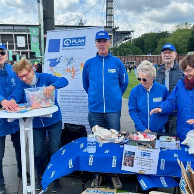 Sechs ältere Erwachsene in blauen Jacken und Hüten mit dem Logo von Plan International stehen an einem Stand im Freien. Auf dem Tisch liegen Broschüren, Spendendosen und Materialien. Im Hintergrund sind ein Stadion und eine Menschenmenge zu sehen, was auf eine Veranstaltung oder Spendenaktion hindeutet.
