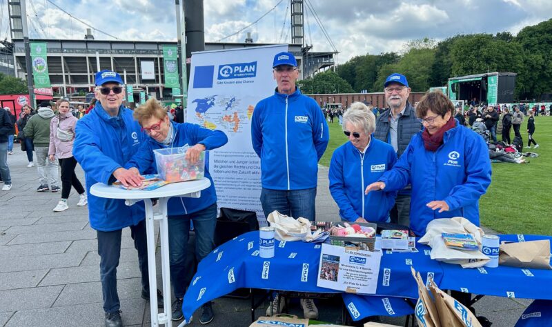 Sechs ältere Erwachsene in blauen Jacken und Hüten mit dem Logo von Plan International stehen an einem Stand im Freien. Auf dem Tisch liegen Broschüren, Spendendosen und Materialien. Im Hintergrund sind ein Stadion und eine Menschenmenge zu sehen, was auf eine Veranstaltung oder Spendenaktion hindeutet.