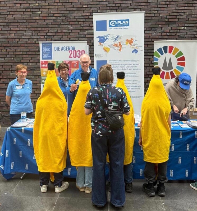 Vier Personen in Bananenkostümen stehen vor einem mit einem blauen Tuch bedeckten Tisch mit Broschüren. Hinter dem Tisch lächeln fünf Personen in blauen Hemden. An der Backsteinmauer im Hintergrund sind Informationsplakate und Banner angebracht.