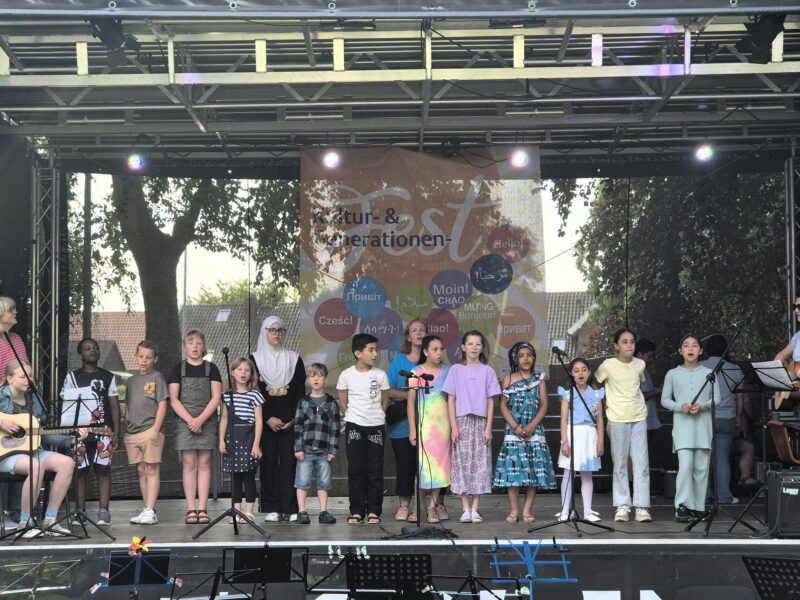 Eine bunte Gruppe von Kindern steht auf einer Freiluftbühne und singt in Mikrofone. Hinter ihnen hängt ein Transparent mit der Aufschrift „Kultur & Generationen Fest“. Zu beiden Seiten sitzen einige Erwachsene mit Instrumenten. Im Hintergrund sind Bäume und Gebäude zu sehen.
