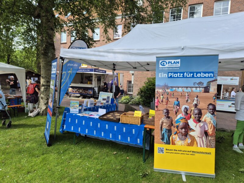 Freiluftmesse mit weißen Zelten auf einer Rasenfläche; an einem Stand sind blaue Banner und Materialien von PLAN International zu sehen. Ein großes Plakat zeigt lächelnde Kinder und einen Text auf Deutsch. Im Hintergrund schlendern Menschen an den Ständen vorbei, im Hintergrund sind Bäume und ein Backsteingebäude zu sehen.