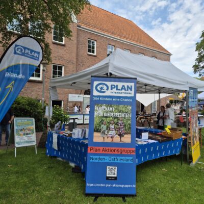 Vor einem Backsteingebäude steht auf einer Rasenfläche ein Stand von Plan International mit Bannern und Schildern. Der Stand wirbt für Kinderrechte und präsentiert Flugblätter, Broschüren sowie ein großes Schild mit der Aufschrift „Plan Aktionsgruppe Norden-Ostfriesland“. Am Stand sind Menschen anwesend.