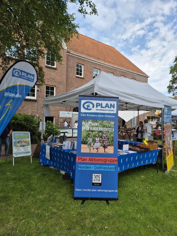 Vor einem Backsteingebäude steht auf einer Rasenfläche ein Stand von Plan International mit Bannern und Schildern. Der Stand wirbt für Kinderrechte und präsentiert Flugblätter, Broschüren sowie ein großes Schild mit der Aufschrift „Plan Aktionsgruppe Norden-Ostfriesland“. Am Stand sind Menschen anwesend.