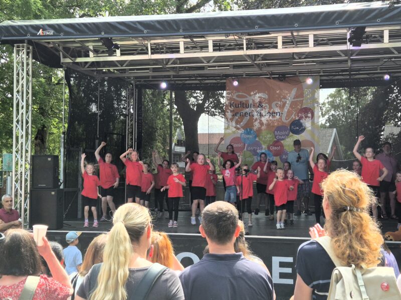 Eine Gruppe von Kindern in einheitlichen roten T-Shirts führt auf einer Freiluftbühne einen choreografierten Tanz auf. Das Publikum schaut zu, einige klatschen. Die Kulisse ist mit bunten Grafiken und deutschen Texten gestaltet. Im Hintergrund sind Bäume und Häuser im Tageslicht zu sehen.