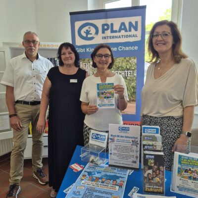 Vier lächelnde Erwachsene stehen hinter einem blauen Tisch, auf dem Informationsmaterial und Broschüren von Plan International ausliegen. Ein Banner mit dem Logo ist zu sehen. Auf dem Tisch liegen Faltblätter, Flyer, eine Spendenbox und Stifte – in einem hellen Raum mit großen Fenstern.