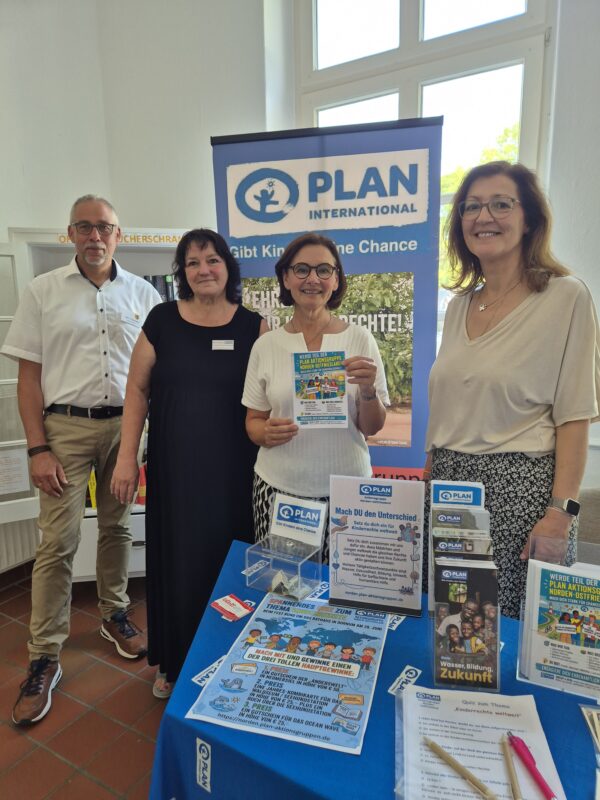 Vier lächelnde Erwachsene stehen hinter einem blauen Tisch, auf dem Informationsmaterial und Broschüren von Plan International ausliegen. Ein Banner mit dem Logo ist zu sehen. Auf dem Tisch liegen Faltblätter, Flyer, eine Spendenbox und Stifte – in einem hellen Raum mit großen Fenstern.
