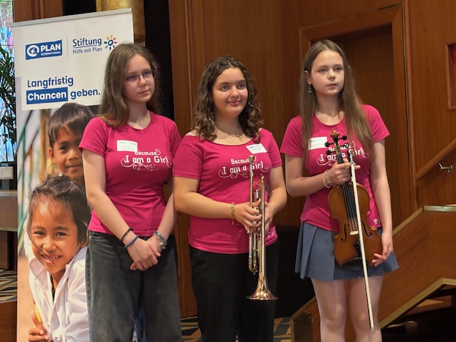 Drei Mädchen stehen in einem Raum und tragen einheitliche rosa T-Shirts mit der Aufschrift „I am a Girl“. Eine hält eine Trompete in der Hand, eine andere eine Geige. Im Hintergrund ist ein Banner mit Kindergesichtern und einem deutschen Text über das Thema „Chancen bieten“ zu sehen. Die Kulisse wirkt formell und ist gut ausgeleuchtet.