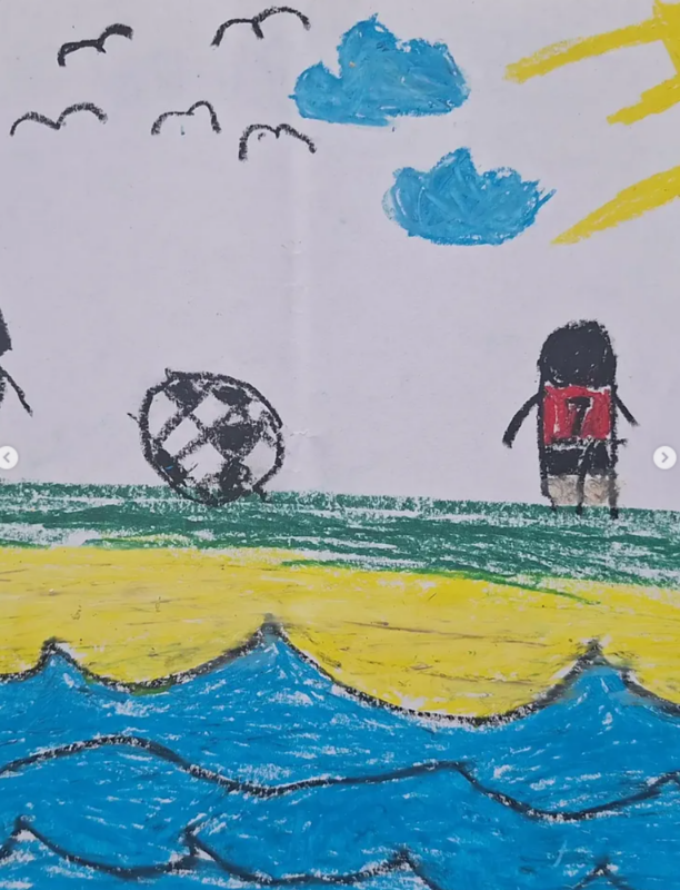 Die Kreidezeichnung eines Kindes zeigt einen Fußballspieler in einem roten Trikot mit der Aufschrift "17" auf dem Rücken, der neben einem schwarz-weißen Fußball auf dem Rasen steht. Blaue Wellen, gelber Sand, Vögel, Wolken und eine Sonne füllen den Hintergrund und bilden eine Strandszene.