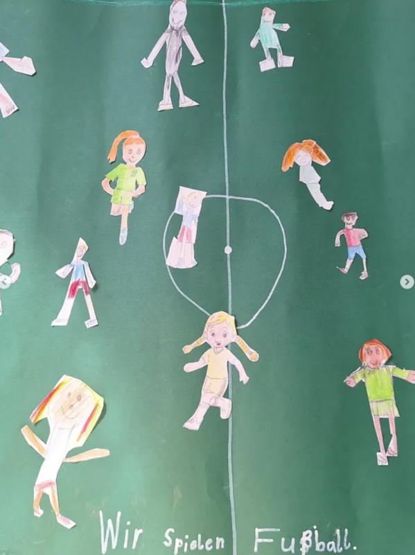 Kinderzeichnung eines Fußballplatzes mit ausgeschnittenen Papierfiguren von fußballspielenden Kindern. Die Figuren sind über das grüne Feld verstreut, einige rennen oder kicken, unten steht in weißer Schrift "Wir spielen Fußball".
