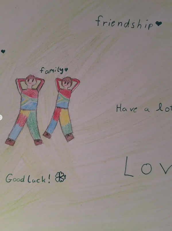 Kinderzeichnung mit zwei Personen in bunten Kleidern, die zusammenstehen und jeweils die Hände hinter dem Kopf verschränken. Der Text um sie herum lautet "Freundschaft", "Familie", "Viel Glück!" mit einem Kleeblatt, "Viel Glück" und "LIEBE". Hellgrüner und gelber Hintergrund.