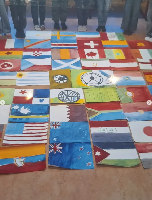 Eine Gruppe von Menschen steht hinter Reihen von handgemalten Nationalflaggen, die auf dem Boden angeordnet sind und verschiedene Länder repräsentieren. Oben sind nur die Beine und Füße der Menschen zu sehen; die Flaggen sind bunt und auf rechteckige Papierbögen gemalt.