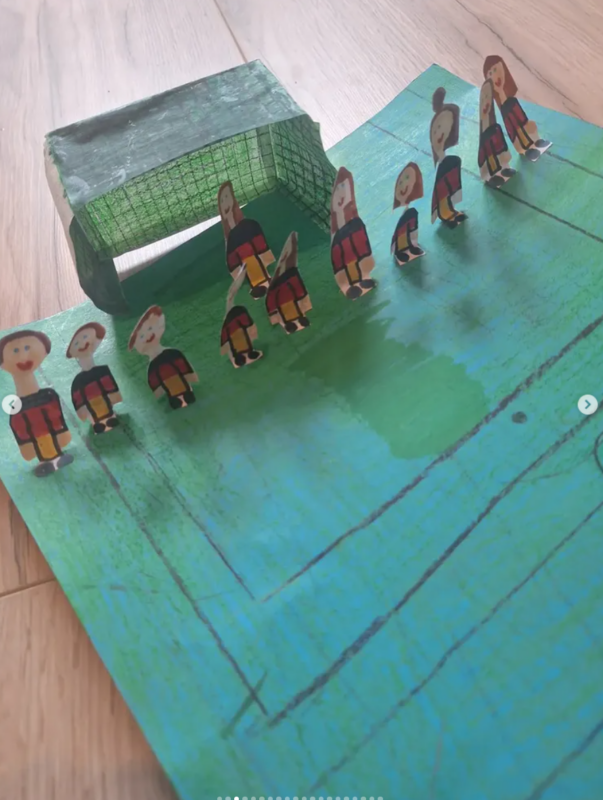 Ein handgefertigtes Fußballfeld mit einem grünen Papptor und elf aus Pappe ausgeschnittenen Spielern in roten und schwarzen Trikots, die alle auf einem gezeichneten grünen Feld mit sichtbaren Spielfeldmarkierungen stehen, das auf einem Holzboden steht.