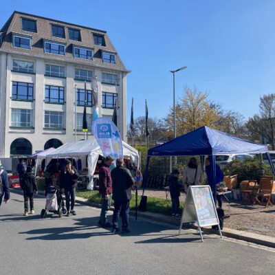 An einem sonnigen Tag versammeln sich Menschen an Ständen und in Zelten im Freien in der Nähe eines großen, modernen Gebäudes. Einige stehen, andere sitzen an Holztischen, und ein Kinderwagen ist zu sehen. Ein blaues Banner und ein Schild weisen auf eine Veranstaltung hin, im Hintergrund sind Bäume mit frühlingshaftem Laub zu sehen.