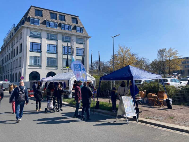 An einem sonnigen Tag versammeln sich Menschen an Ständen und in Zelten im Freien in der Nähe eines großen, modernen Gebäudes. Einige stehen, andere sitzen an Holztischen, und ein Kinderwagen ist zu sehen. Ein blaues Banner und ein Schild weisen auf eine Veranstaltung hin, im Hintergrund sind Bäume mit frühlingshaftem Laub zu sehen.