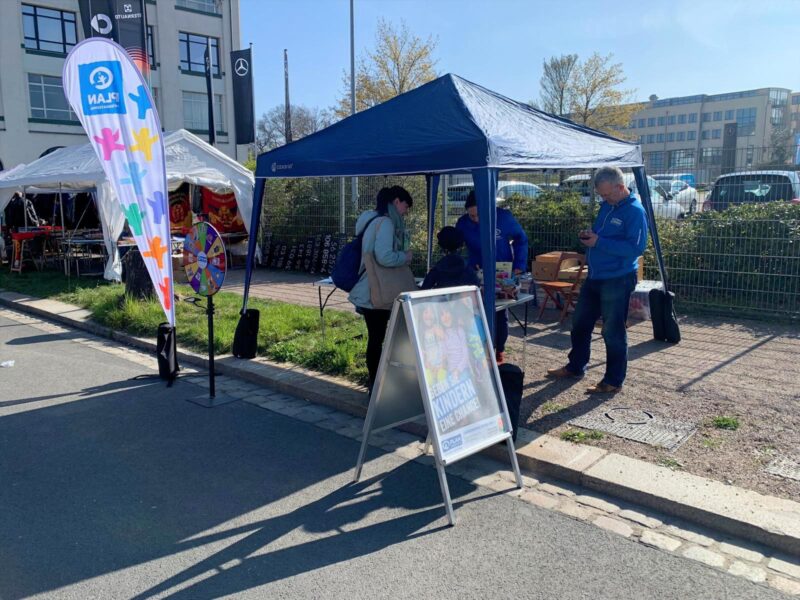 An einer sonnigen Straße steht ein kleiner Stand im Freien mit einem blauen Vordach. Unter dem Zelt unterhalten sich vier Personen. Eine bunte Standfahne und ein Aufsteller machen auf die Veranstaltung aufmerksam. Im Hintergrund sind nahegelegene Gebäude und Grünflächen zu sehen.
