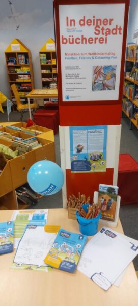 In einer Bibliothek steht ein rot-weißer Ständer mit der Aufschrift "In deiner Stadtbücherei". Darunter liegen Arbeitsblätter, Buntstifte, ein blauer Luftballon und Broschüren auf einem Tisch. Im Hintergrund sind die Regale mit Kinderbüchern gefüllt.