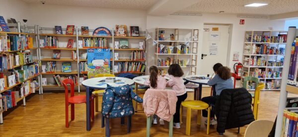 Eine Kinderbibliothek mit bunten Stühlen und runden Tischen. Drei Kinder sitzen da, malen oder lesen, ihre Mäntel sind über die Stühle drapiert. An den Wänden stehen Bücherregale, gefüllt mit Büchern und Spielen. Der Boden ist aus Holz, und die Atmosphäre wirkt gemütlich und einladend.