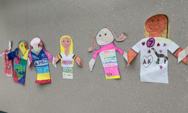 Fünf bunte, handgezeichnete Papierpuppen sind an eine gesprenkelte Wand geklebt. Jede Puppe ist anders dekoriert, mit leuchtenden Farben, Kleidungsmustern, Schrift und Symbolen, die einzigartige Designs und Persönlichkeiten darstellen.