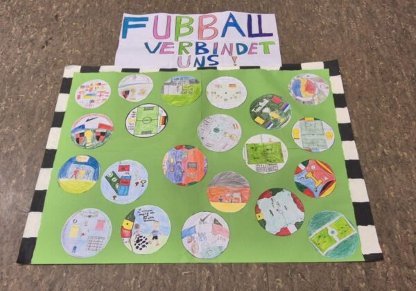 Ein grünes Plakat zeigt 16 handgezeichnete Kreise mit bunten Bildern zum Thema Fußball. Oben steht ein weißes Schild mit der Aufschrift "Fubball verbindet uns!" in Blau, Grün und Lila, was auf Deutsch "Fußball verbindet uns! Das Plakat steht auf einem braunen Boden.